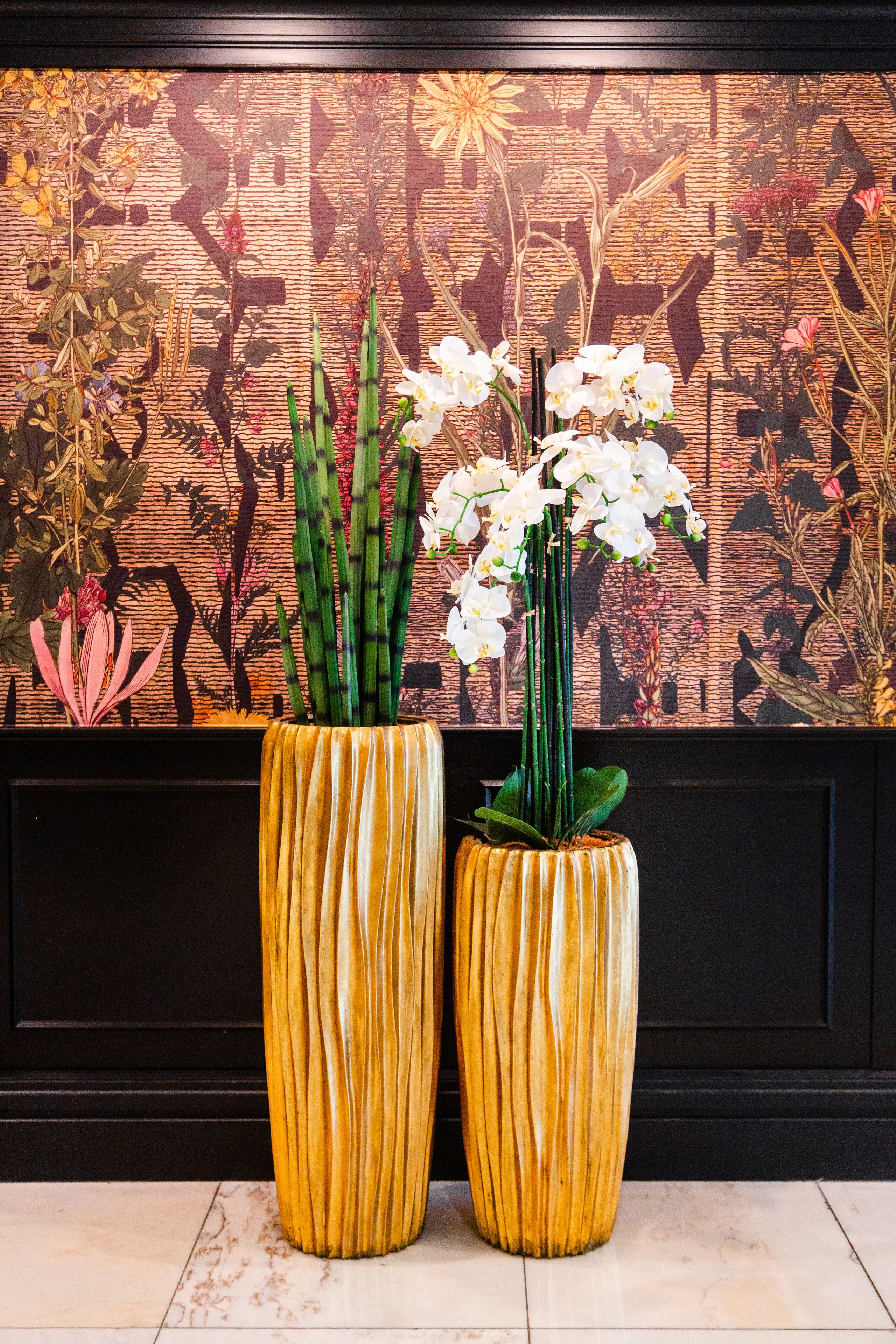 Two tall, yellow, ribbed vases with white orchids on a tiled floor against a patterned wall.