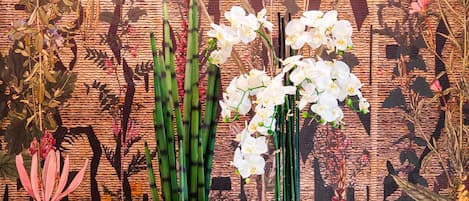 Dois vasos altos e amarelos com nervuras, adornados com orquídeas brancas, sobre um chão de azulejos em frente a uma parede estampada.