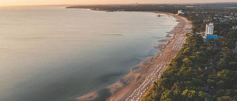 Vue aérienne d’une zone côtière, avec une plage bordée de parasols et quelques bâtiments.