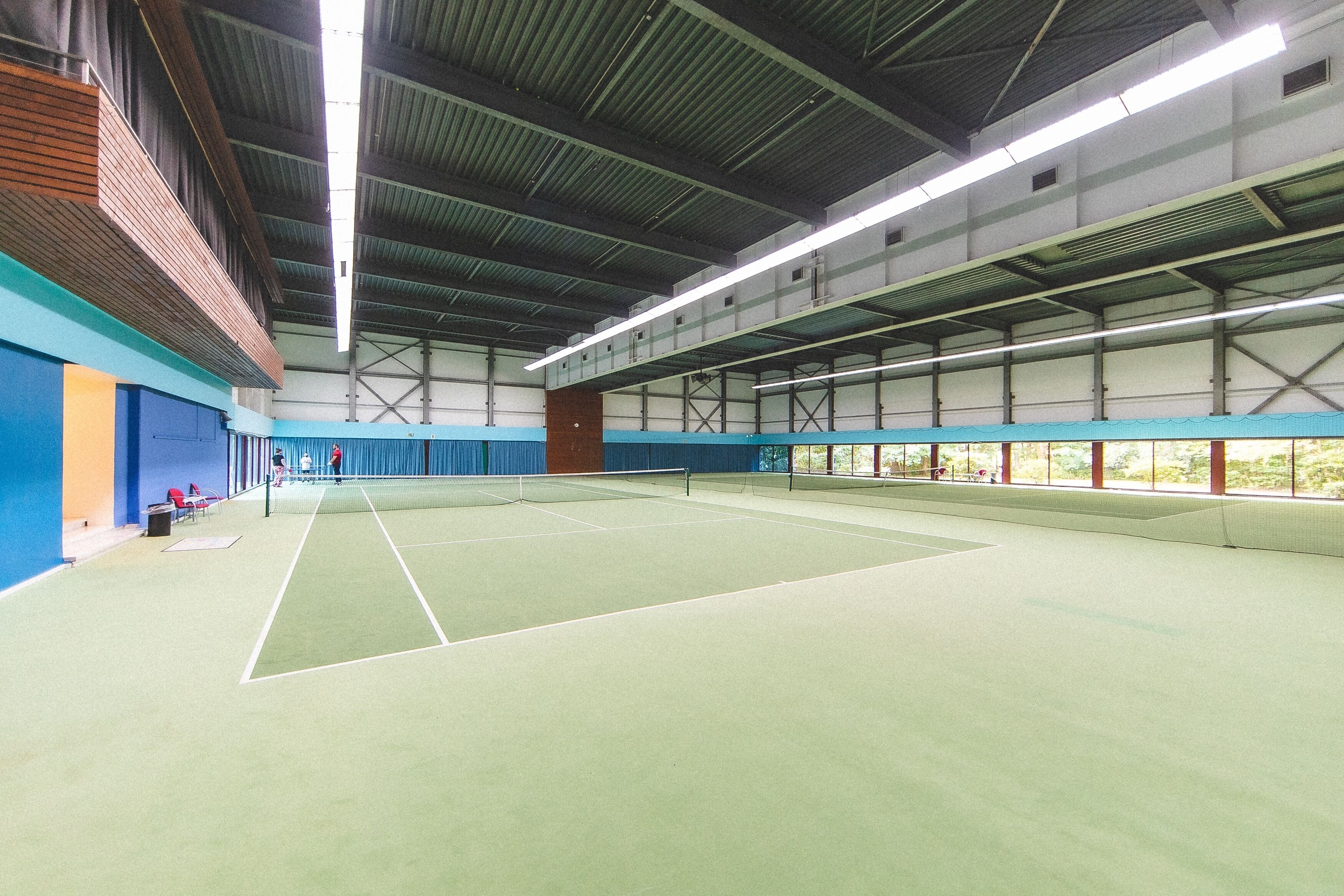 Un court de tennis intérieur doté d’un plafond haut et de poutres métalliques, entouré de murs percés de fenêtres.