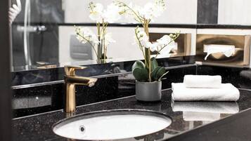 A bathroom with a black countertop, a gold faucet, a white sink, a potted plant, and a towel.