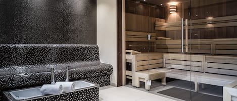 Un baño moderno con un banco de mosaico negro, una sauna con paredes de vidrio y un lavamanos blanco con toallas.