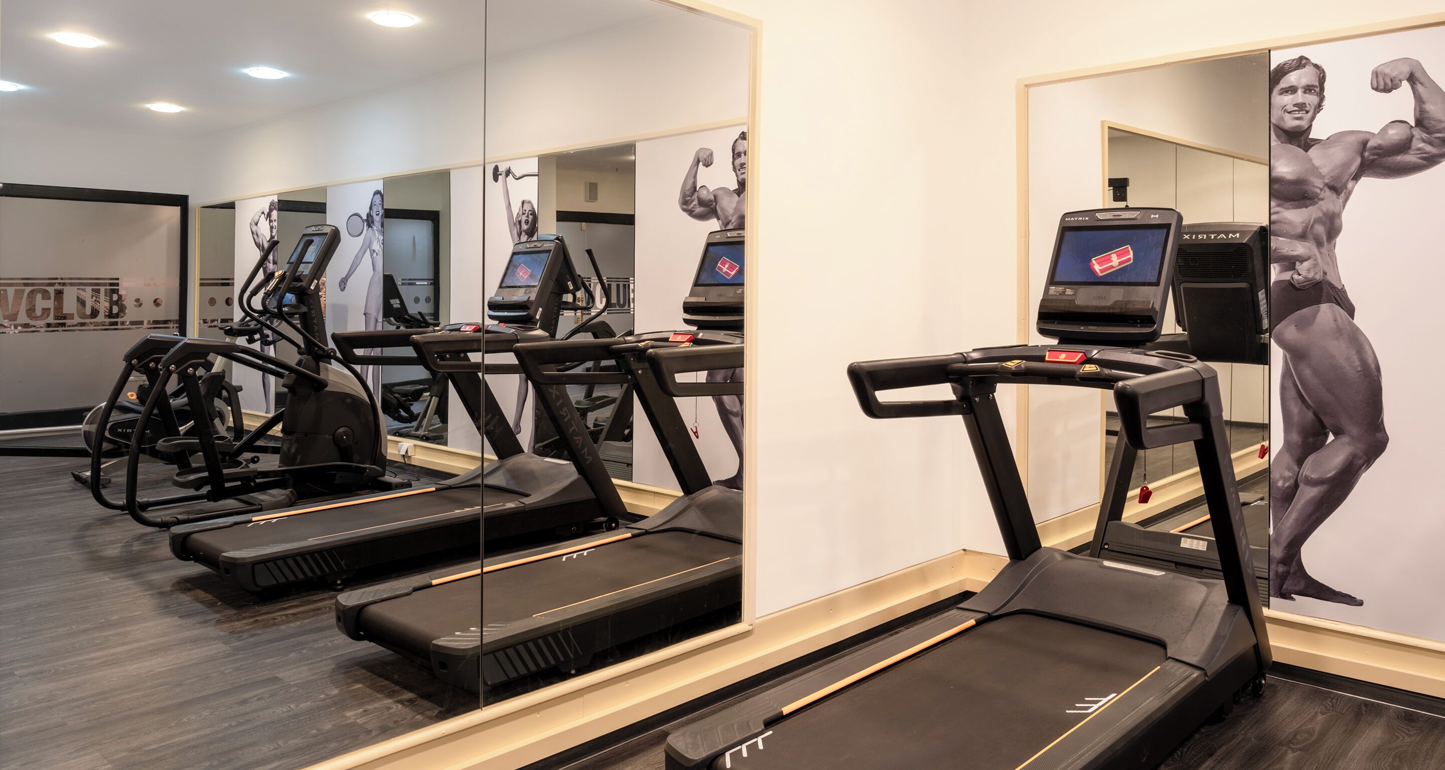 A gym with treadmills, elliptical machines, and a mirrored wall with bodybuilding posters.