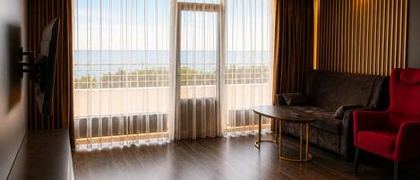 Een moderne hotelkamer met een houten vloer, een bank, een rode fauteuil, een tafel en een grote schuifglazen deur die uitkomt op een balkon met uitzicht.