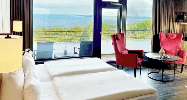 A hotel room with a bed, a red chair, a small table, and a view of the sea. - PLAZA Premium Timmendorfer Strand (Timmendorfer Strand)