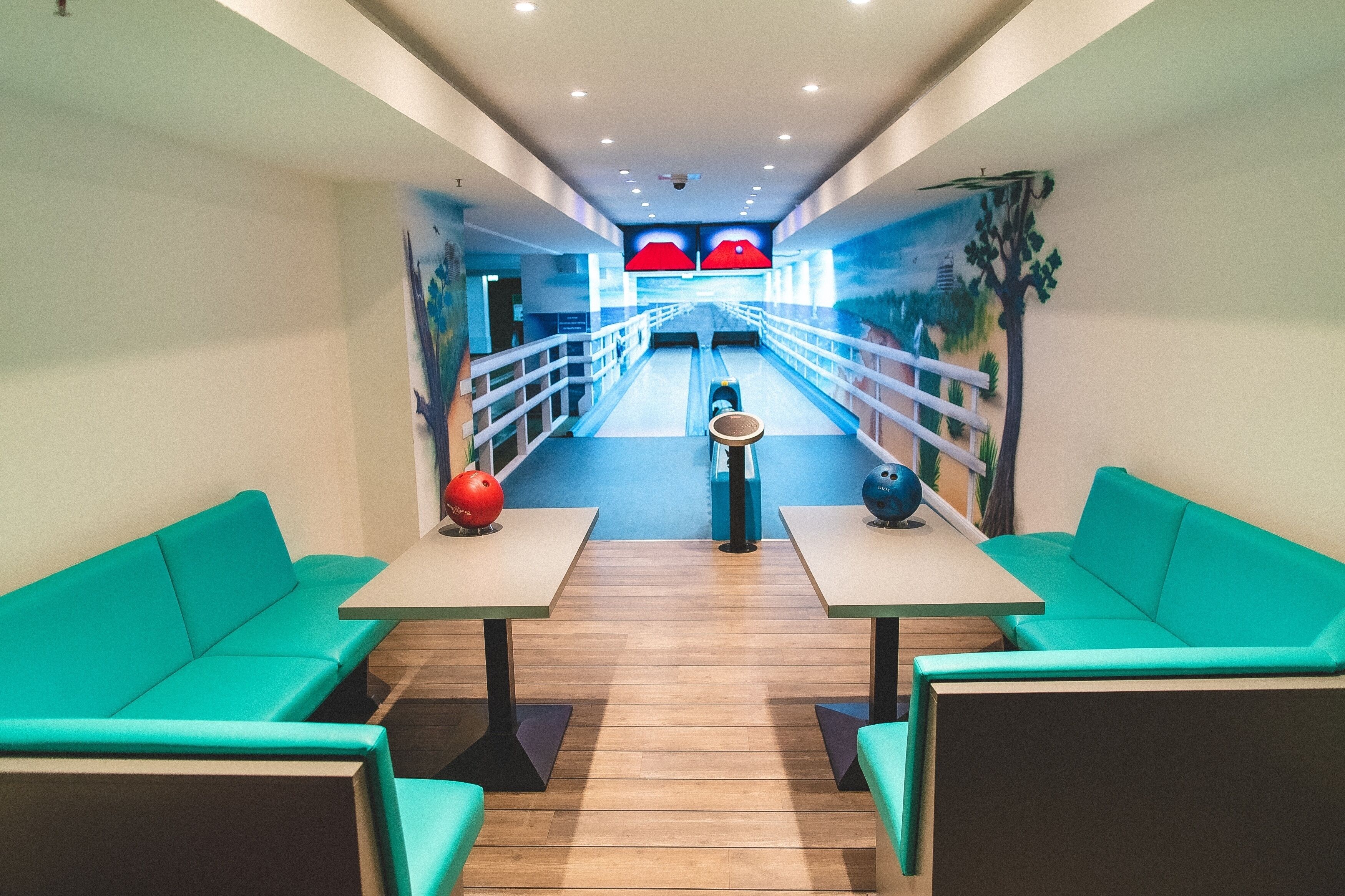 A bowling alley with teal seating, a bowling ball rack, and a mural of a bowling lane.