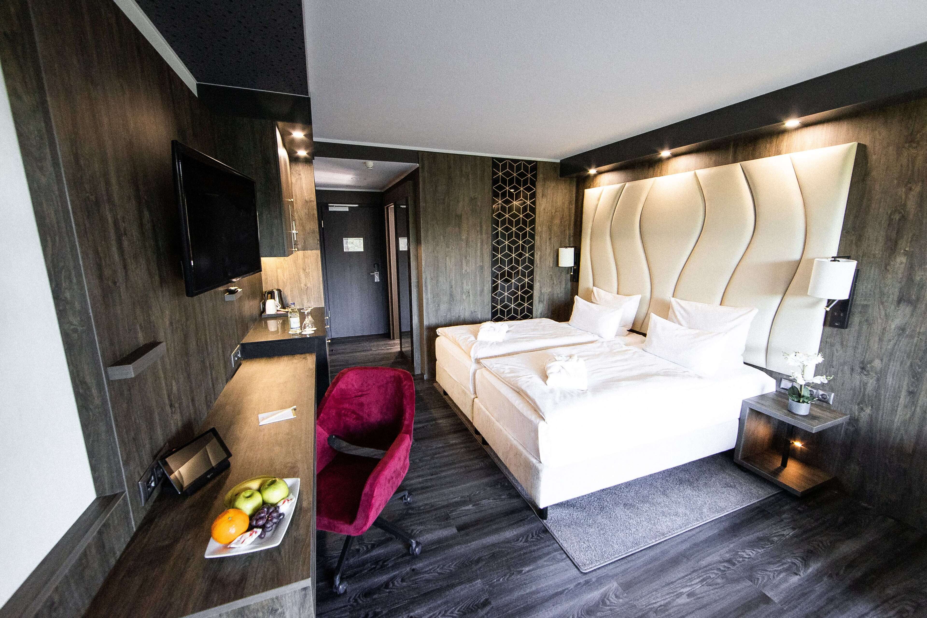 Une chambre d’hôtel moderne avec un grand lit, un téléviseur à écran plat fixé au mur, un fauteuil rouge et une petite table sur laquelle est disposée une assiette de fruits.
