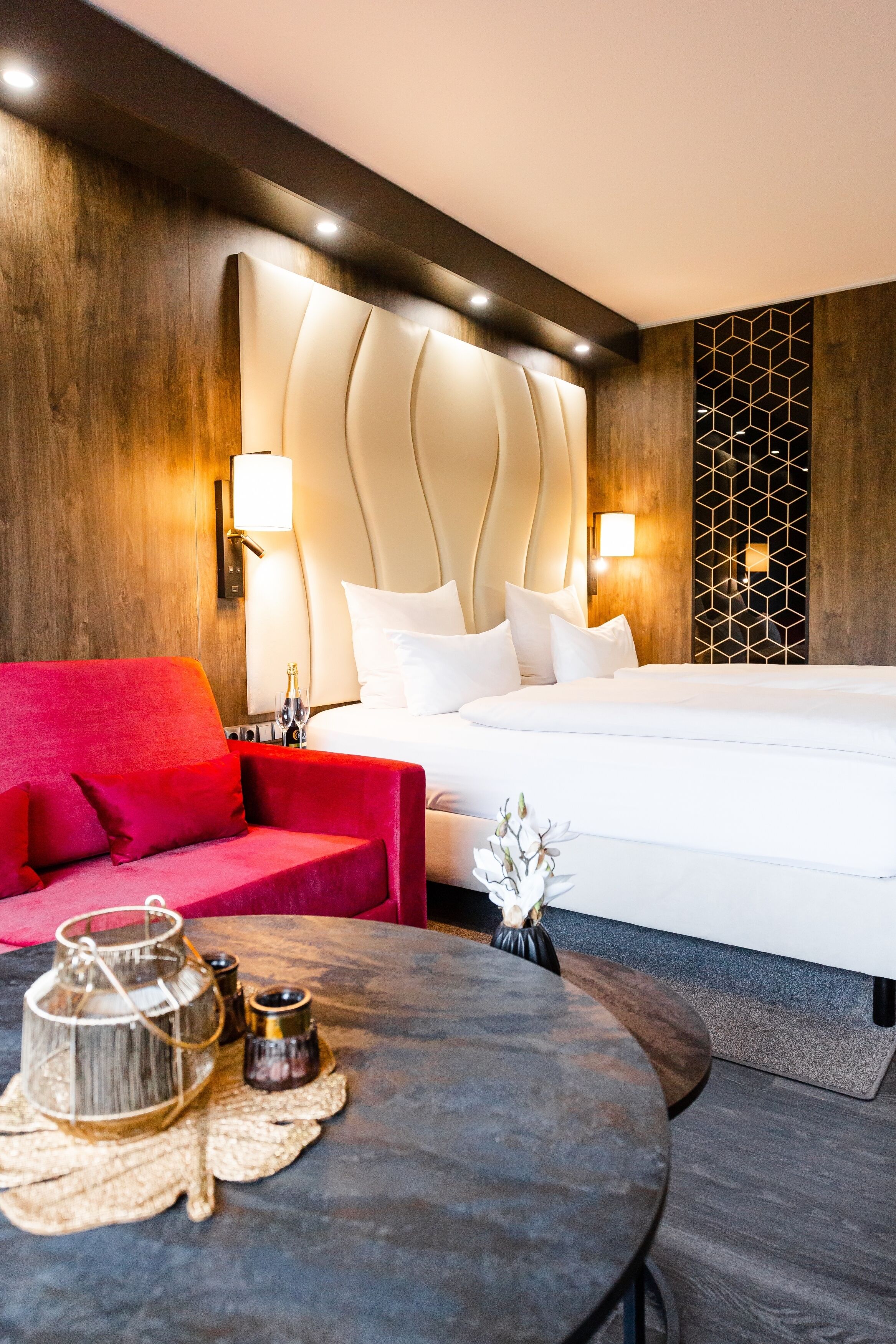A modern hotel room with a large bed, a red sofa, a round table with a candle, and a decorative vase.