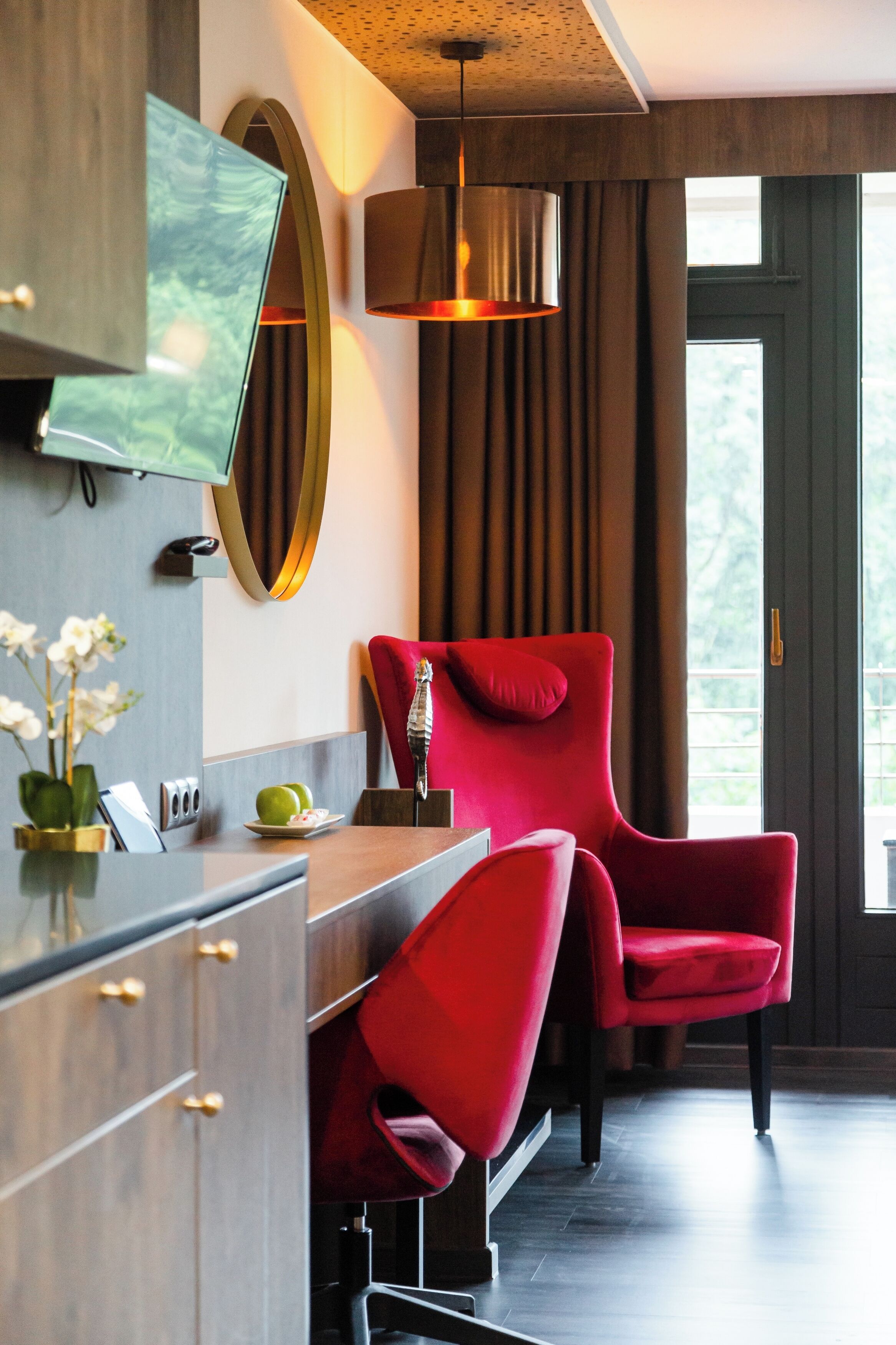 A modern hotel room with a wooden desk, a red chair, a large mirror, and a television mounted on the wall.
