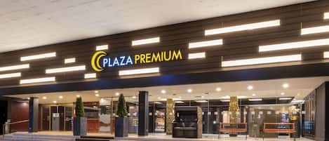 The exterior of Plaza Premium with a modern design, featuring a prominent sign, glass doors, and a seating area with benches.