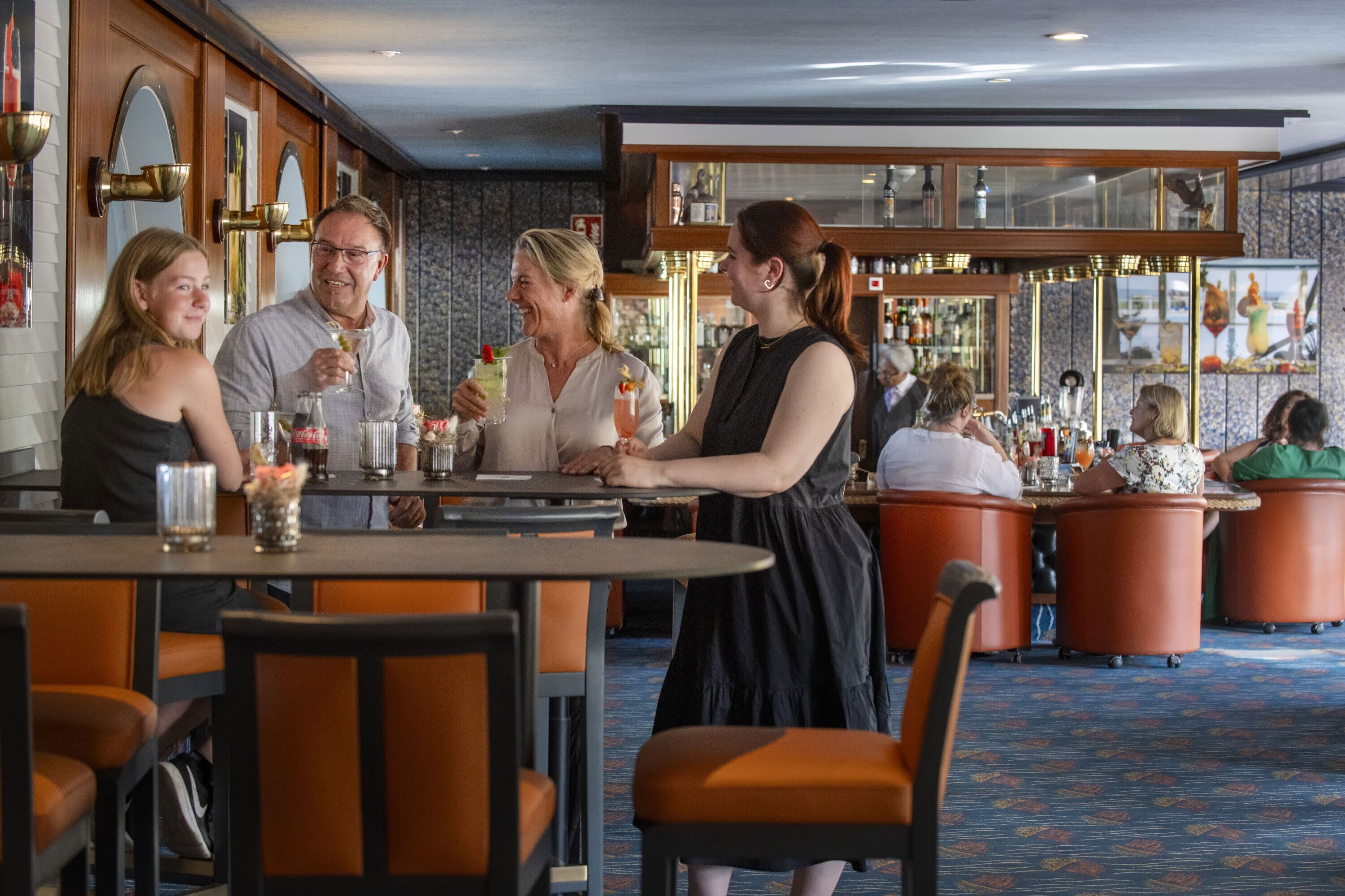 A bar area with round tables and orange chairs, a bartender, and guests seated.