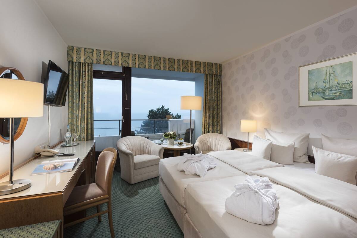 Ein Hotelzimmer mit einem Bett, zwei Sesseln, einem Schreibtisch mit Lampe, einem Fernseher und einem Fenster mit Vorhängen.