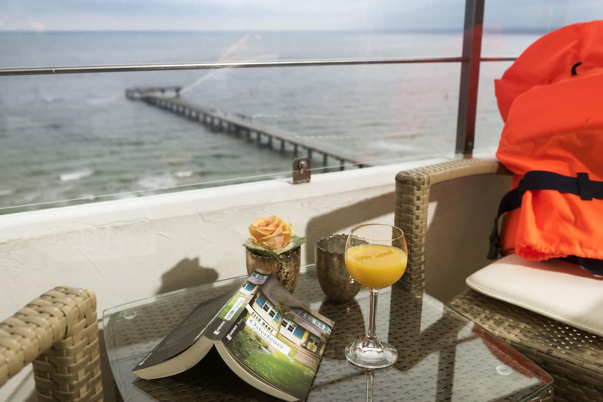 Ein Balkon mit Meerblick, ein Korbstuhl, ein Glastisch mit einer Zeitschrift, ein Glas Orangensaft und eine Rose in einer Vase.