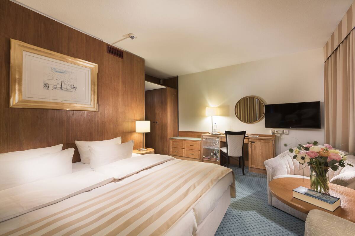 Ein Hotelzimmer mit einem großen Bett, einem Schreibtisch mit Stuhl, einem an der Wand befestigten Fernseher und einer Vase mit Blumen auf einem Beistelltisch.