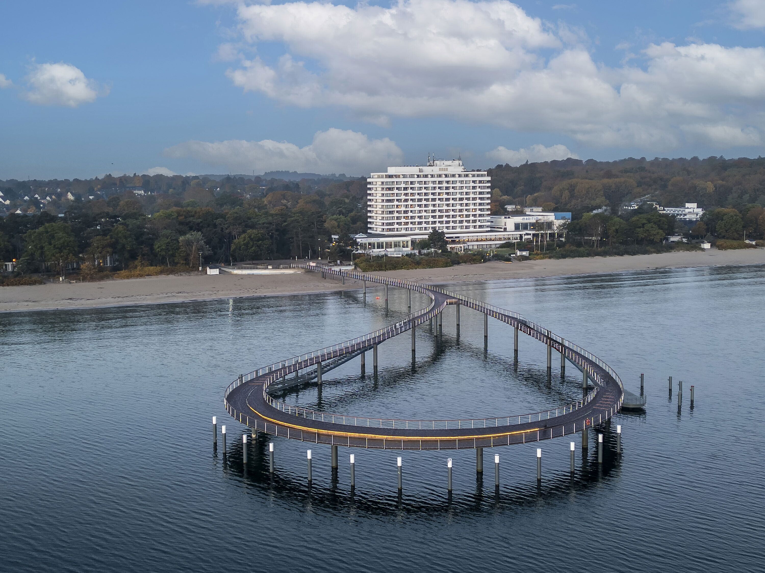 Un hotel frente al agua con un muelle circular que se adentra en el agua, rodeado de un paisaje boscoso y un cielo despejado.