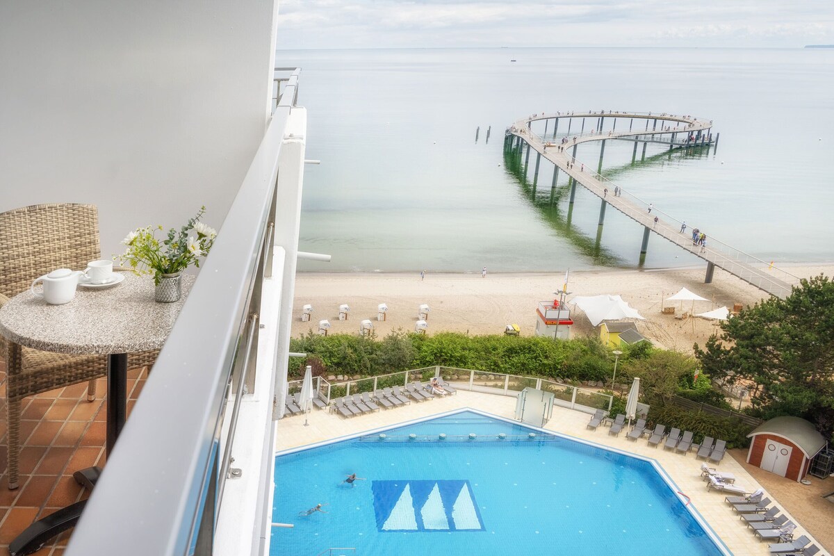 A balcony with a view of a beach, a swimming pool, and a pier extending into the water.