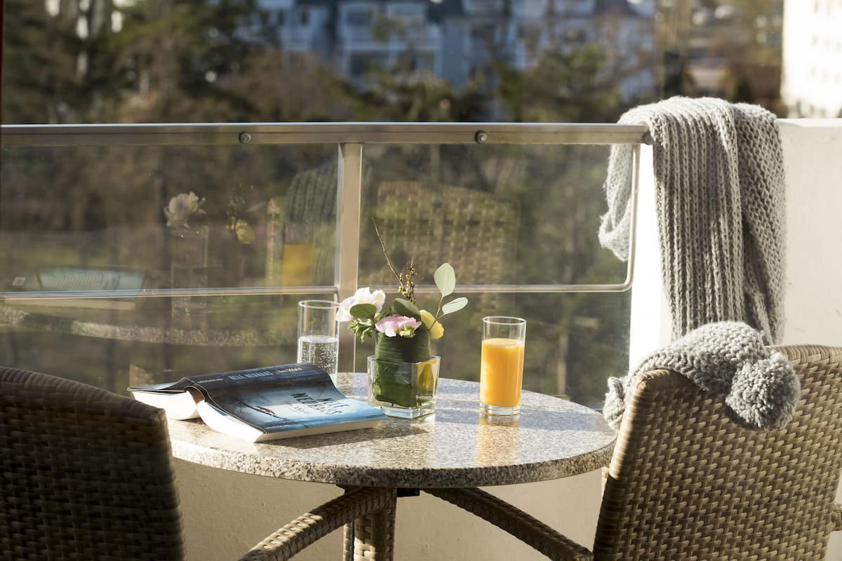 Ein runder Tisch mit einem Buch, einem Glas Orangensaft und einer Blumenvase auf einem Balkon mit Blick auf eine Wohngegend.