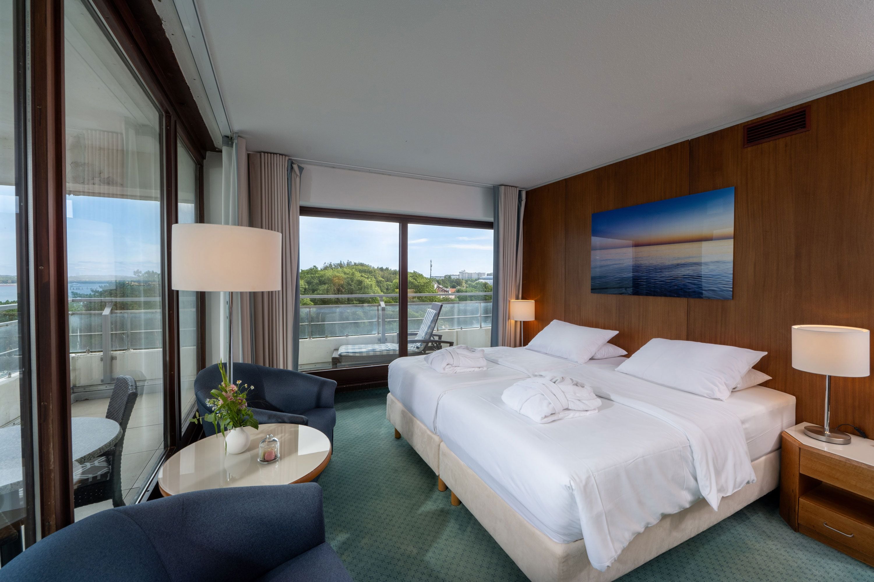 Ein Hotelzimmer mit einem großen Bett, zwei Nachttischlampen, Blick auf die Stadt, einem Balkon mit einem Stuhl und einem kleinen Tisch mit Blumen in einer Vase.