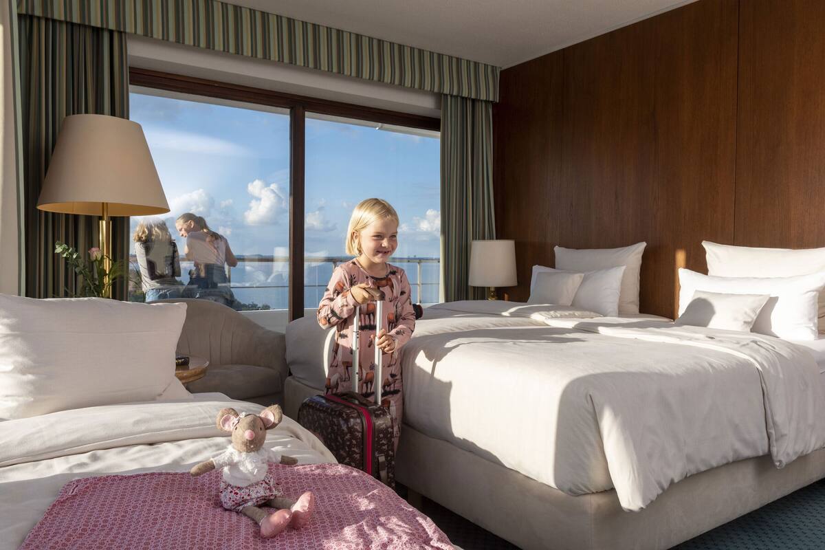 Ein Hotelzimmer mit einem großen Bett, einem kleineren Bett, einer Couch, einer Lampe, einem Fenster mit Vorhängen und Blick aufs Meer.