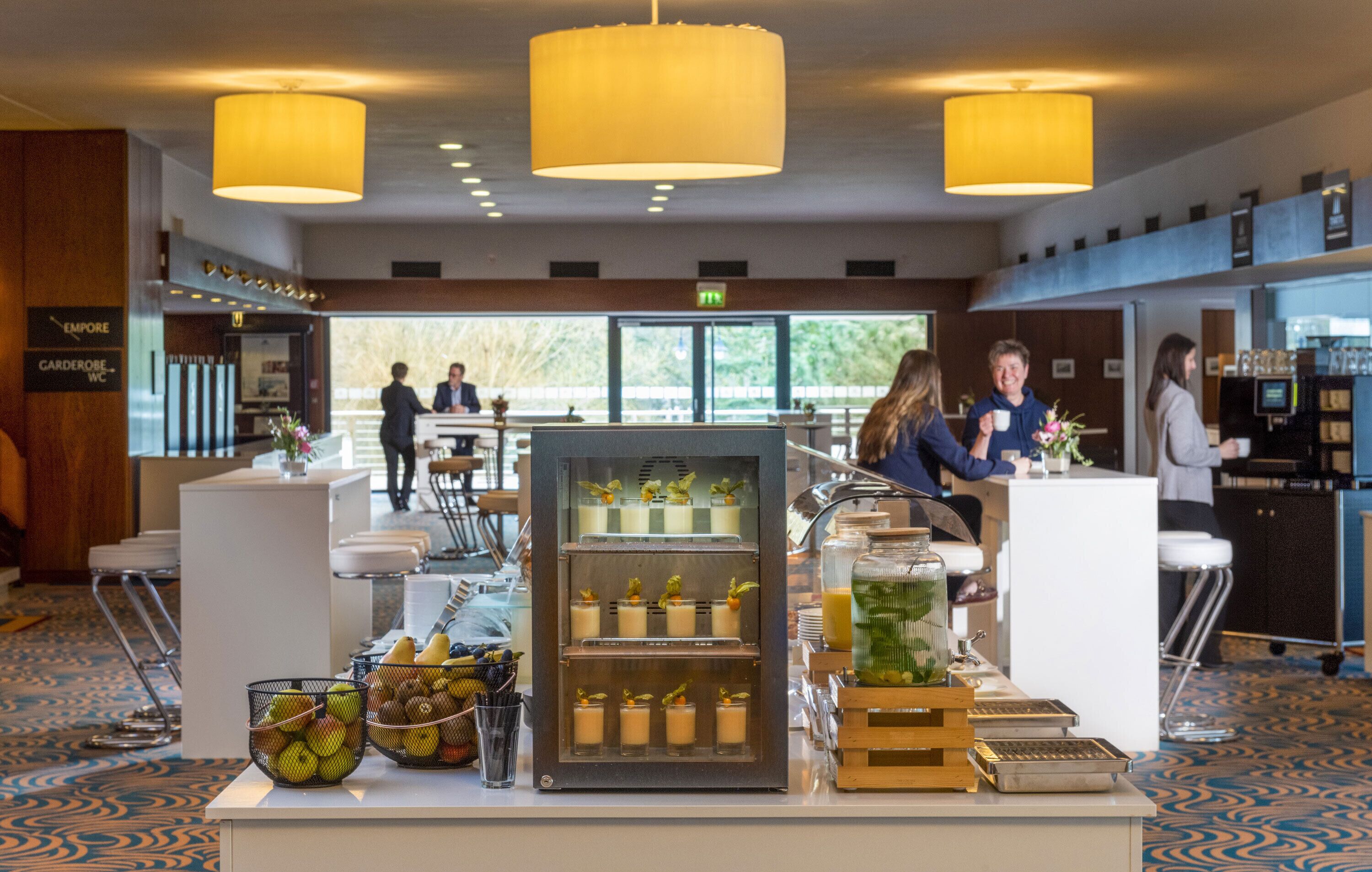 Un hall de hotel con mostrador de recepción, un refrigerador con postres y un área de estar con mesas y sillas.