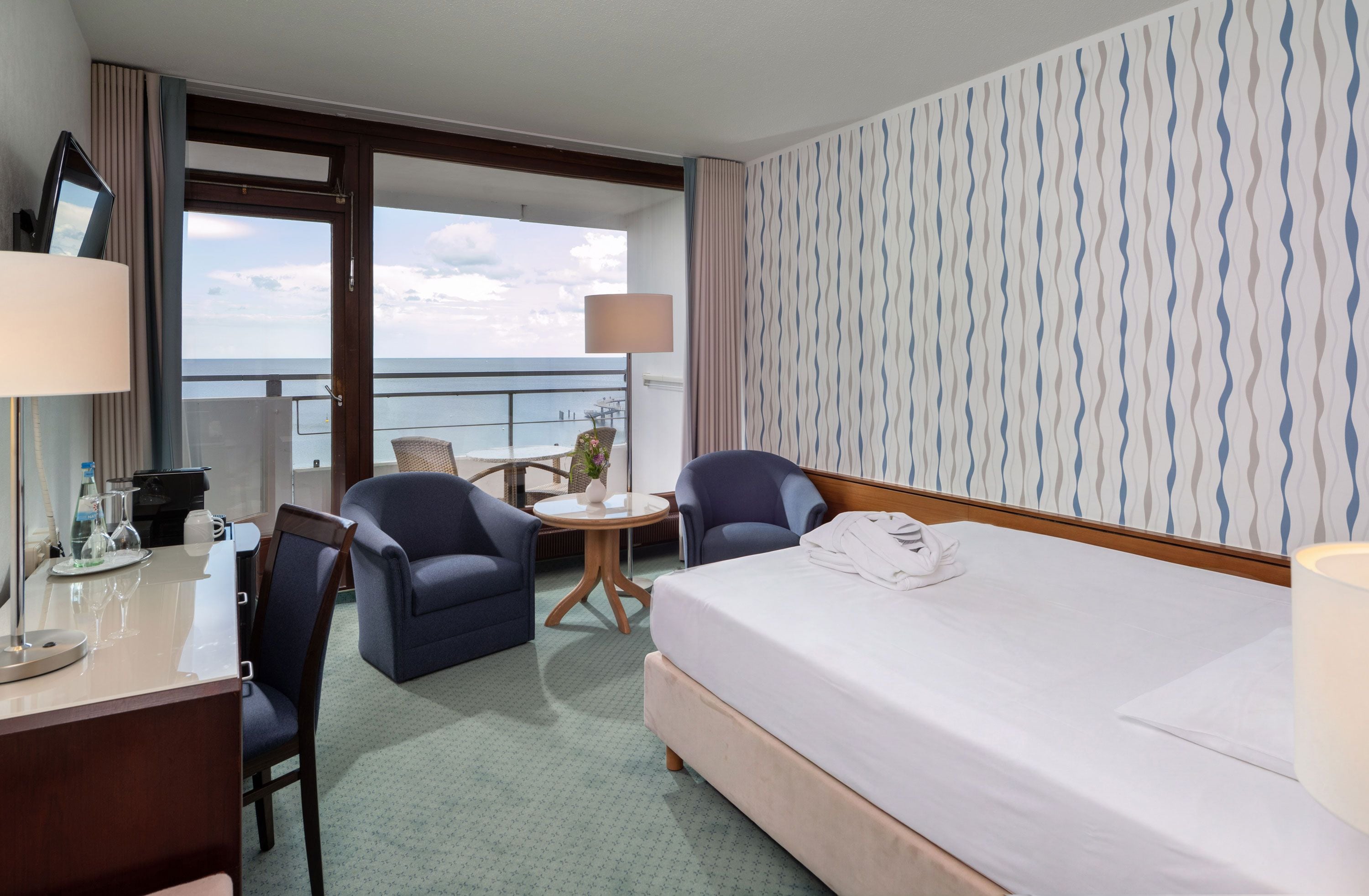 A hotel room with a bed, two armchairs, a small table, a desk with a lamp, and a view of the sea.