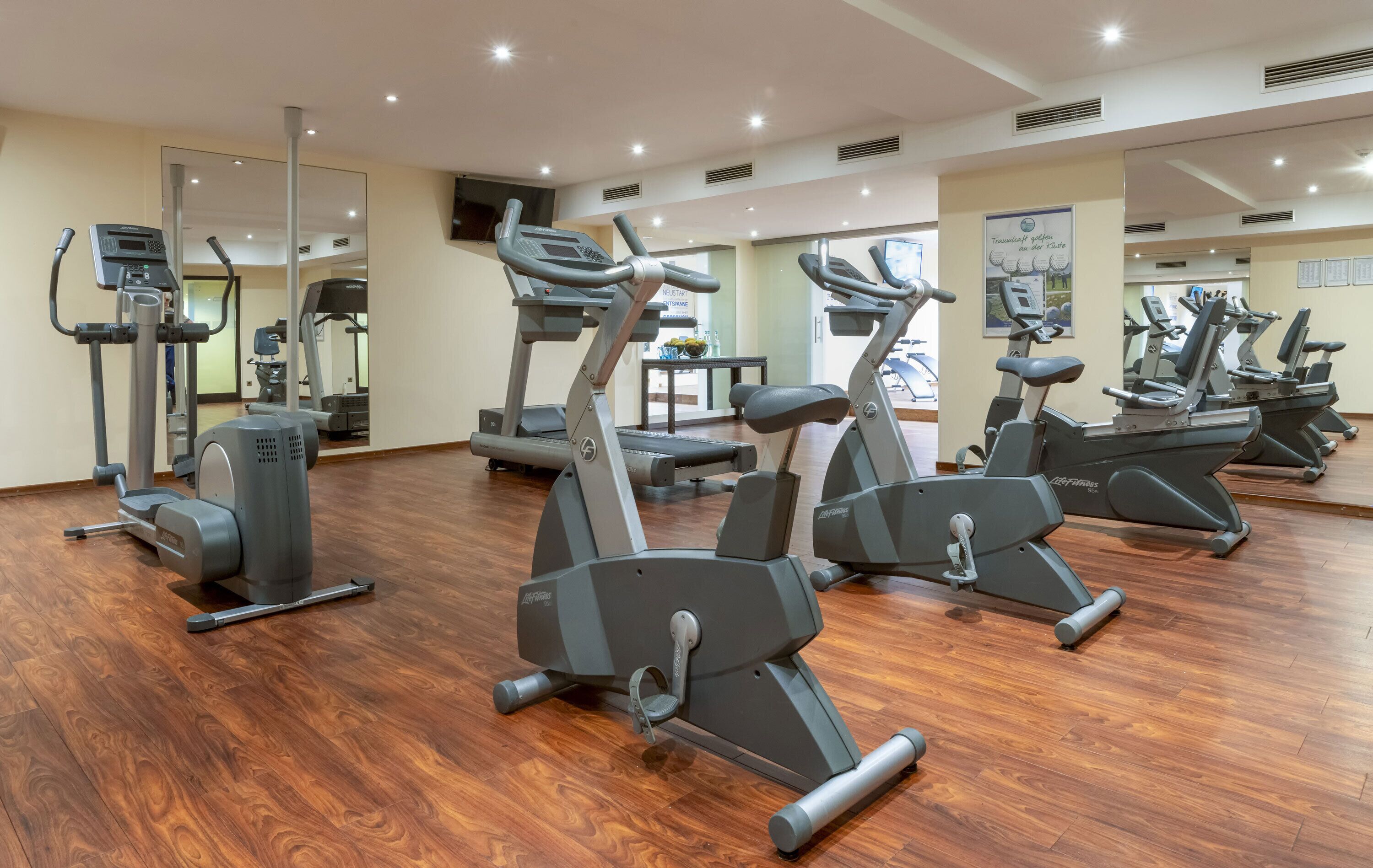 A gym with multiple cardio machines, a mirrored wall, and wooden flooring.