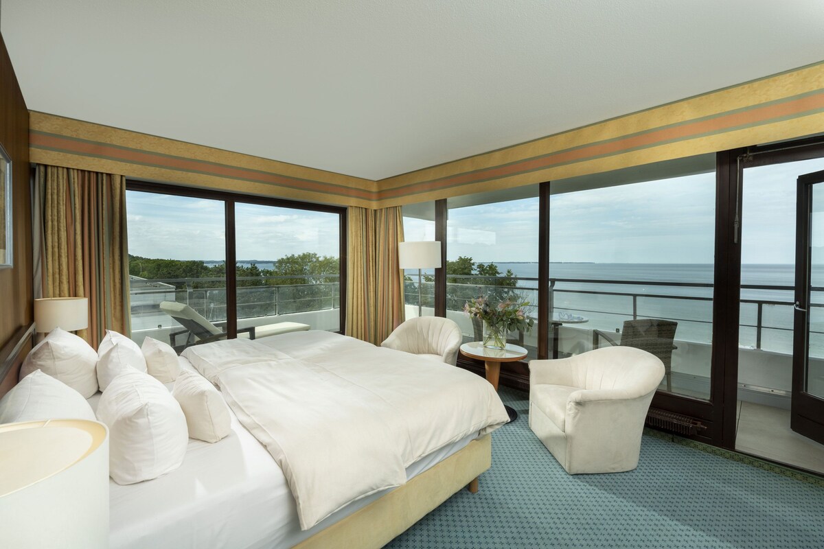 A hotel room with a large bed, a sitting area with a chair and table, and a balcony with a view of the sea.