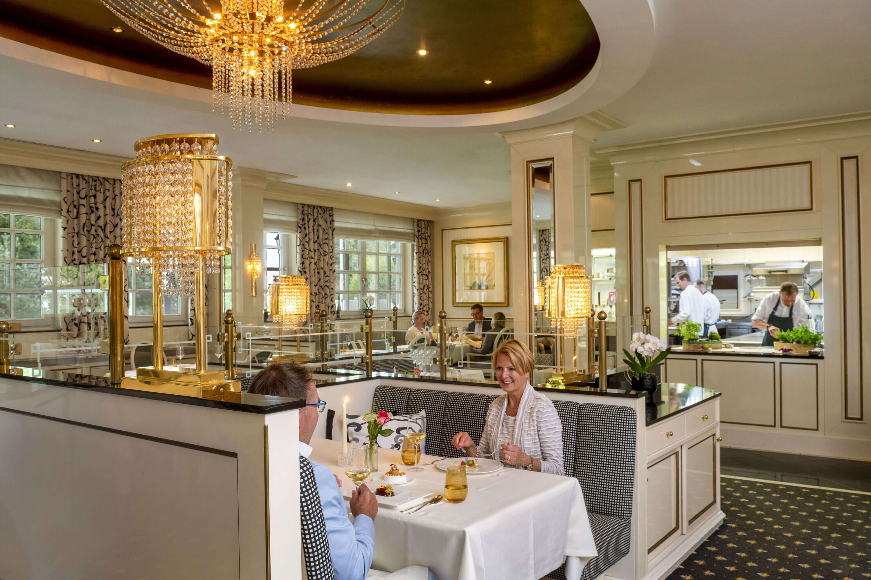 A dining area with a table set for two, a chandelier, and a view of the kitchen in the background.