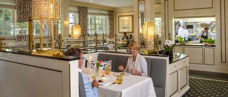 A dining area with a table set for two, a chandelier, and a view of the kitchen in the background.
