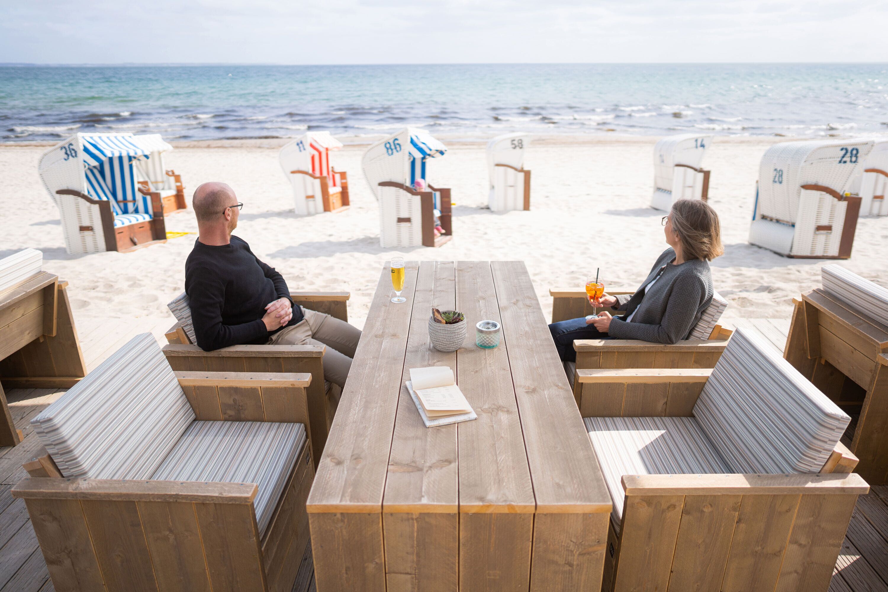 Una terraza de madera con asientos y una mesa, sillas de playa con números, y el océano al fondo.