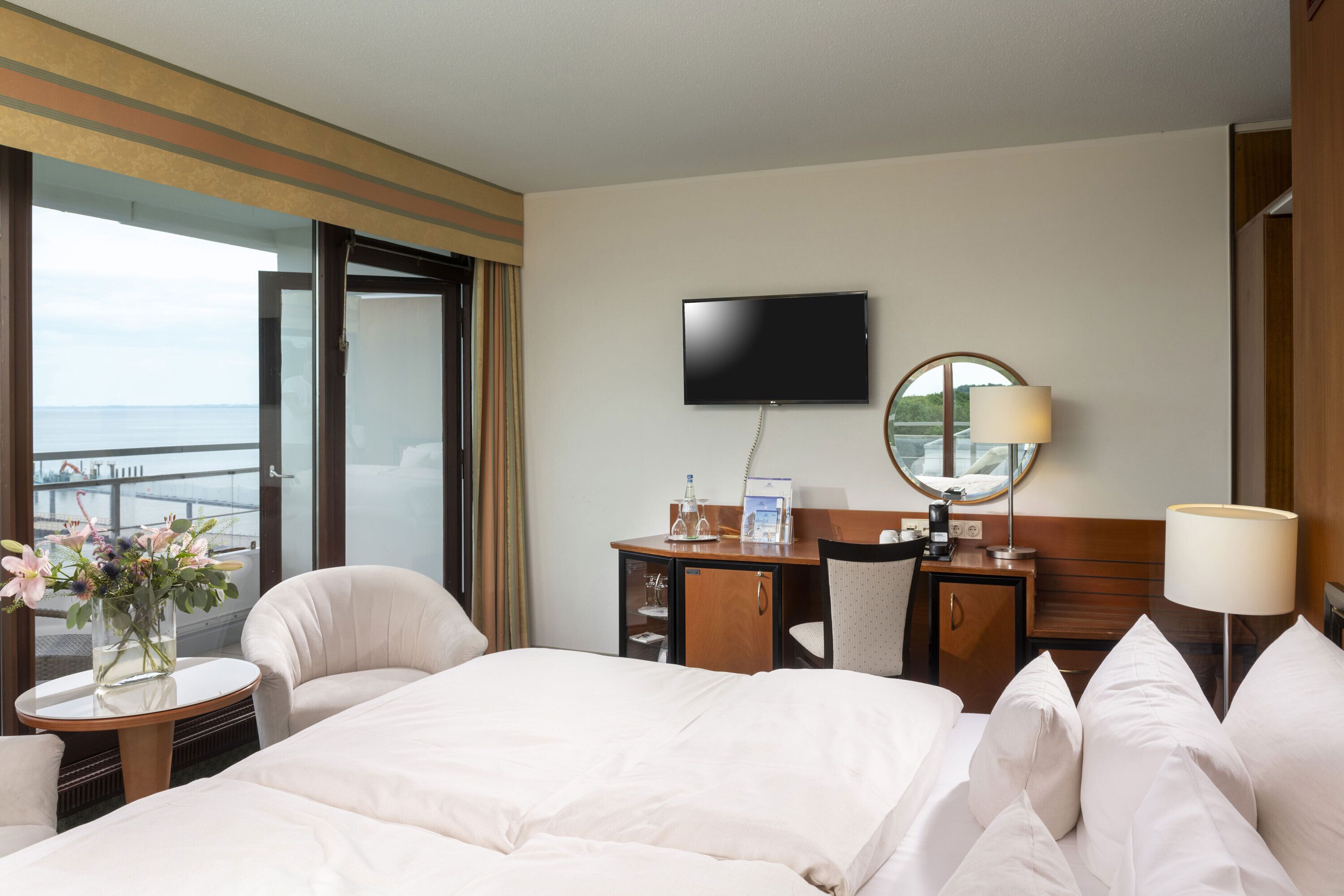 Habitación de hotel con una cama grande, un escritorio con silla, televisión y vista al mar a través de un ventanal amplio.