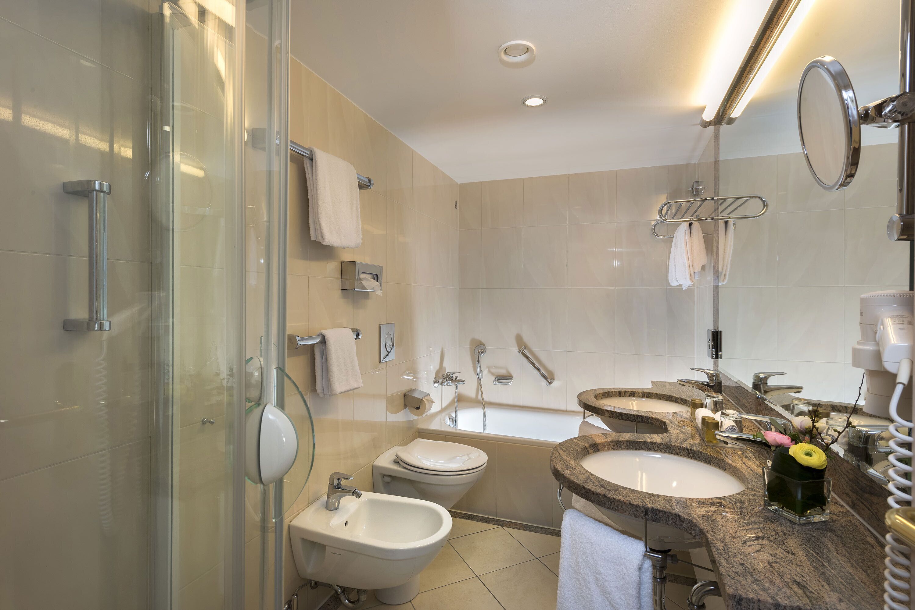 A modern bathroom with a glass shower enclosure, a bidet, a bathtub, and a vanity with two sinks and a mirror.
