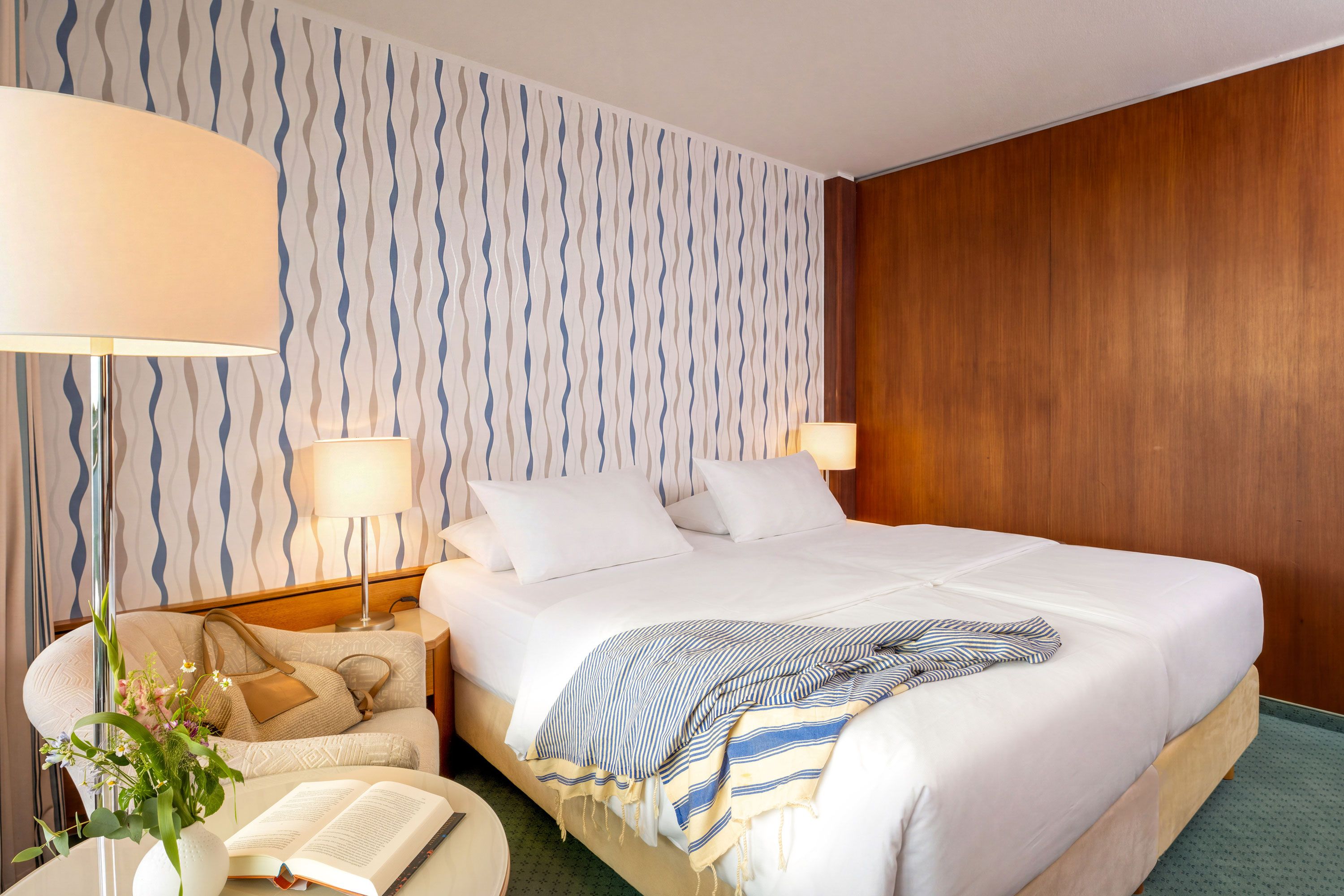 Habitación de hotel con una cama grande, dos lámparas de noche, una mesita de noche con un libro y flores, y una pared con diseño.