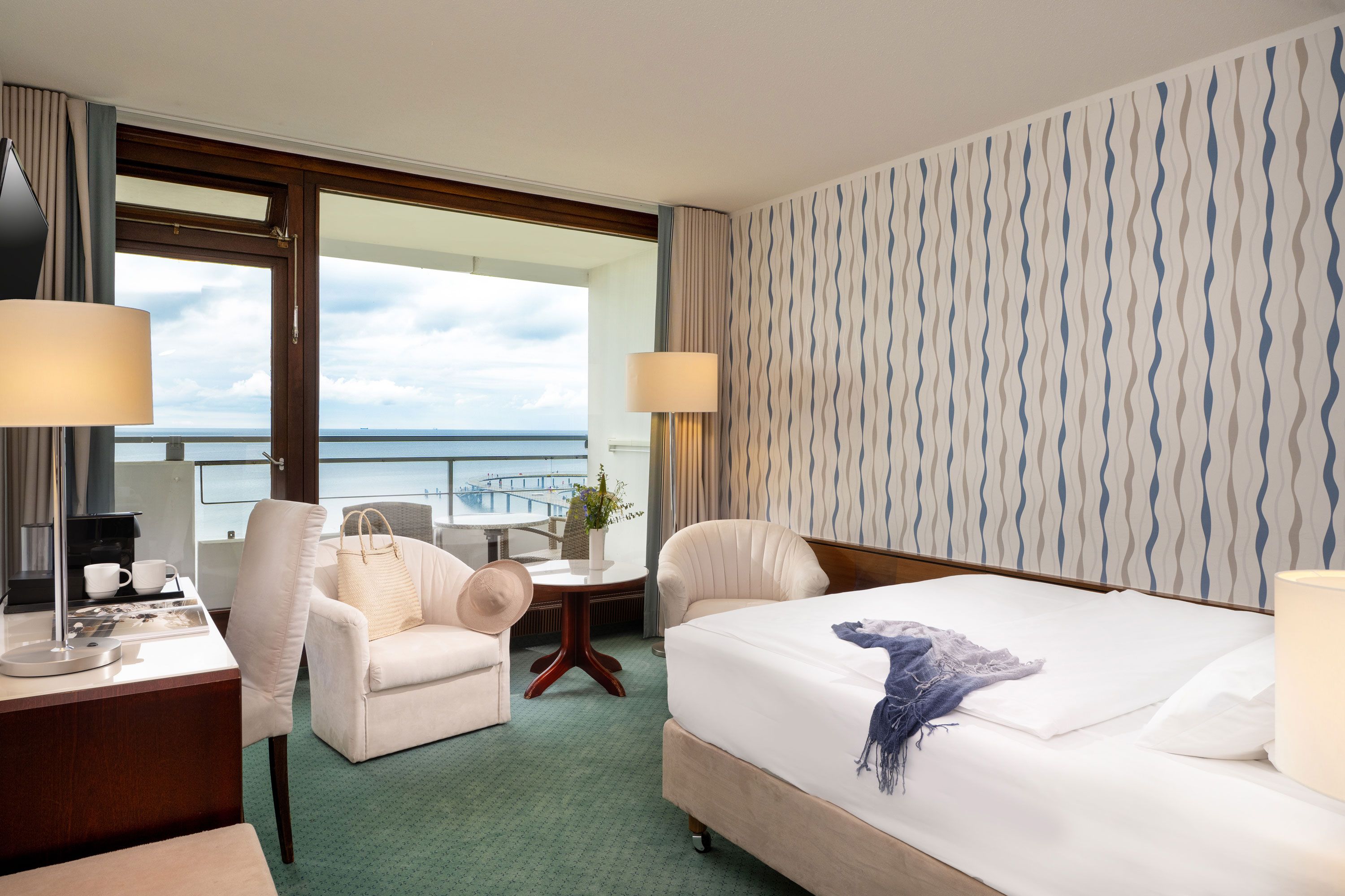 Una camera d'albergo con un letto, due poltrone, una scrivania con una lampada, un tavolino con una pianta e vista sul mare attraverso ampie finestre.