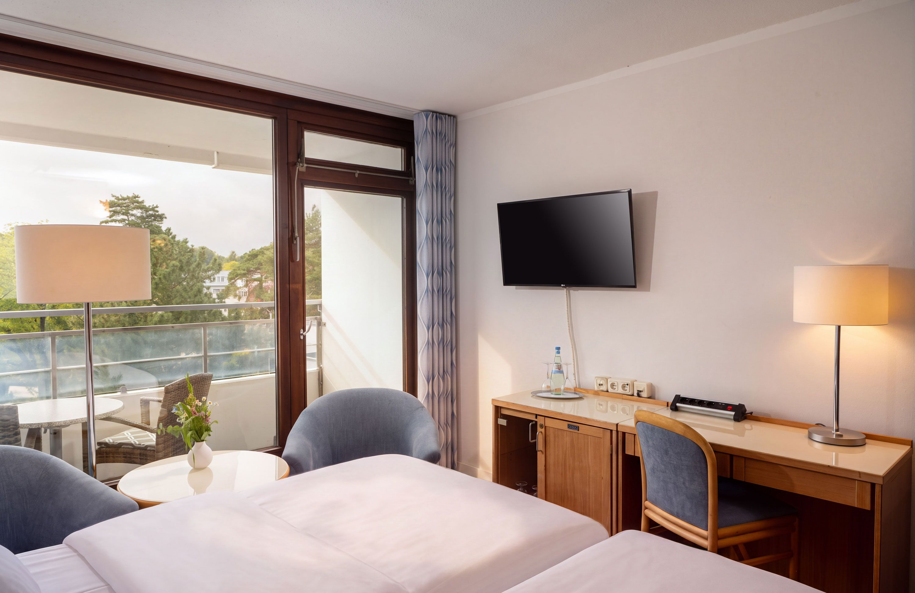 Ein Hotelzimmer mit Bett, Schreibtisch, Sessel, Fernseher und Fenster mit Blick auf Bäume und Gebäude.