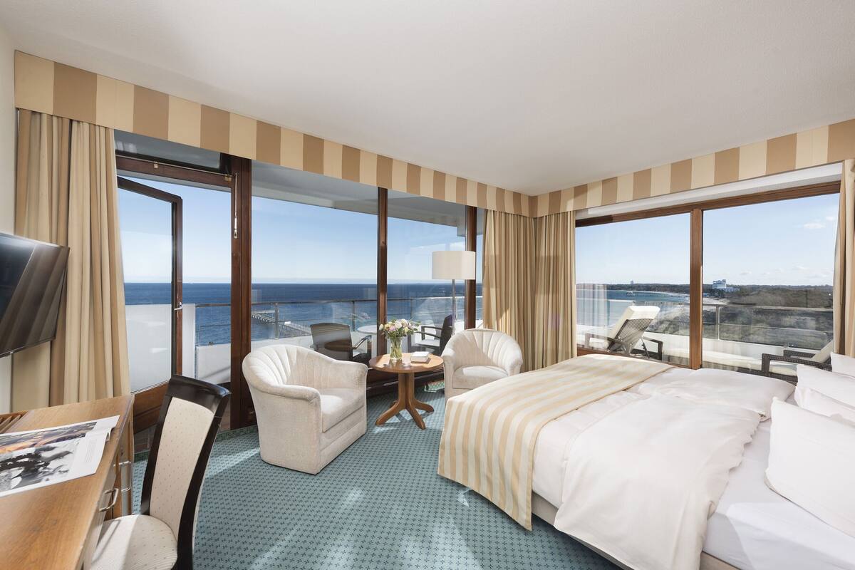 Ein Hotelzimmer mit einem großen Bett, zwei Sesseln, einem kleinen Tisch mit einer Vase und einem Fernseher. Das Zimmer hat große Fenster mit Blick aufs Meer.