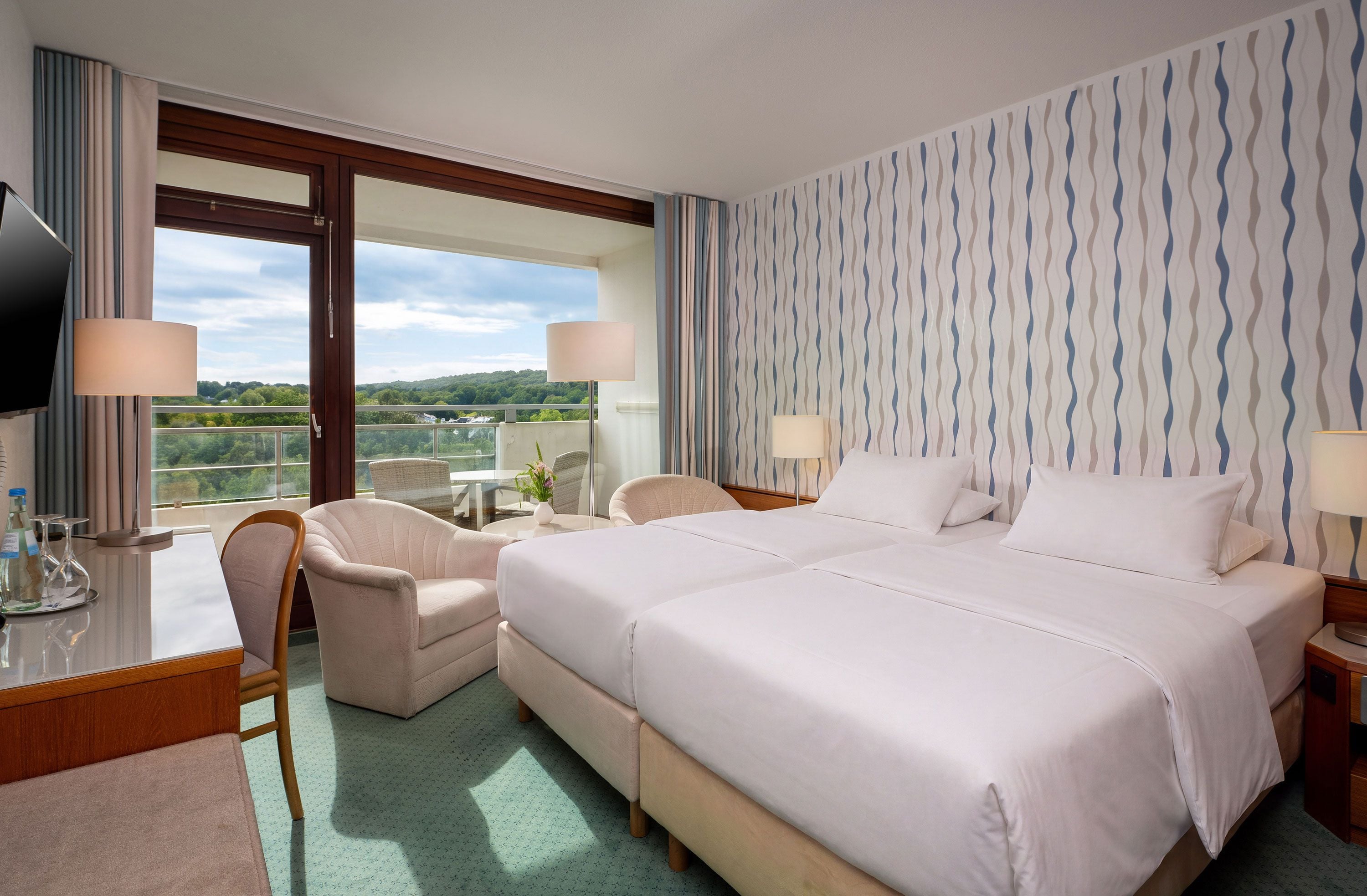 Habitación de hotel con una cama grande, dos mesitas de noche con lámparas, un escritorio con silla, un televisor y un balcón con vistas a zonas verdes.