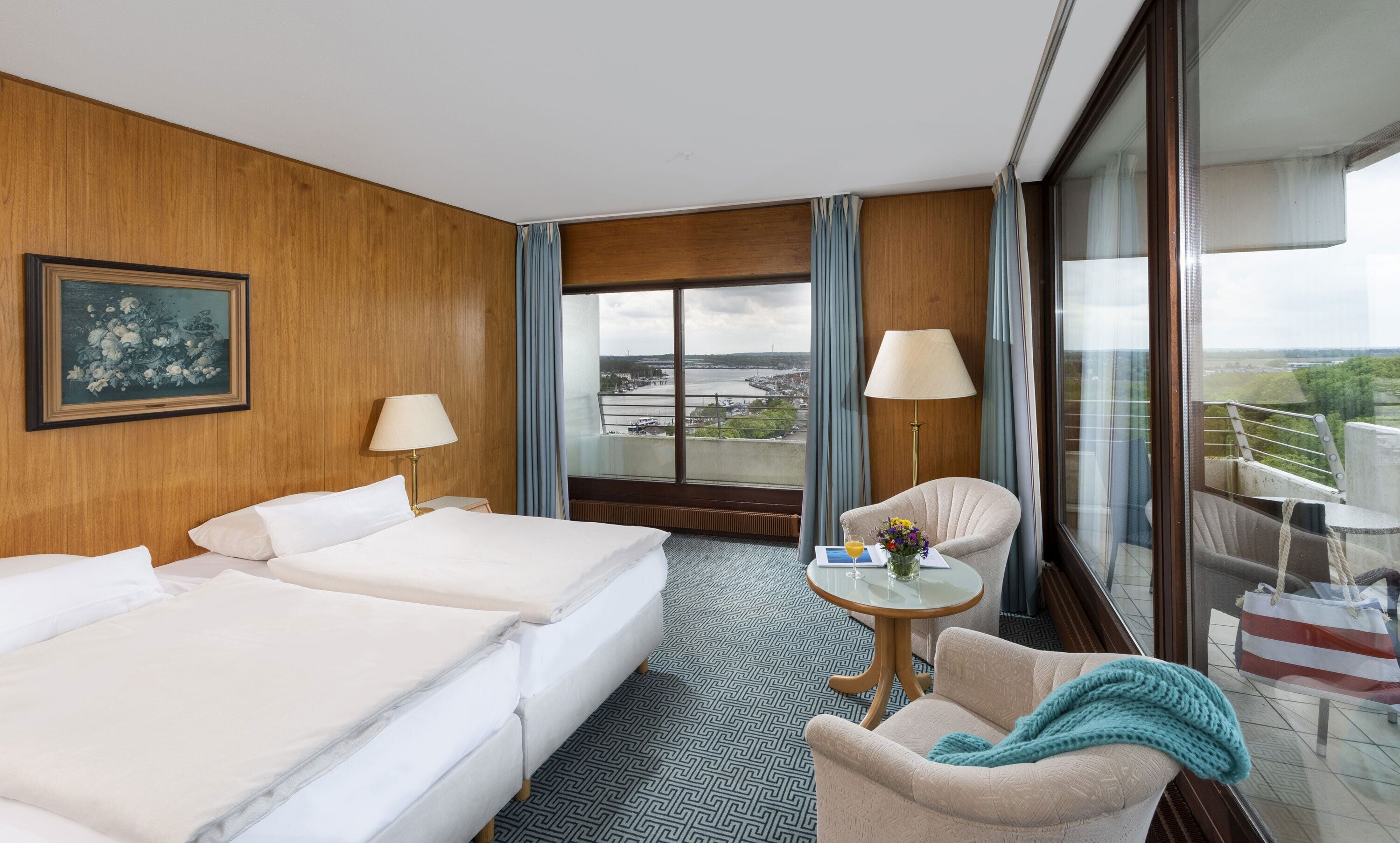 Ein Hotelzimmer mit zwei Betten, einem kleinen Tisch mit Blumen in einer Vase, einem Sessel und einem Balkon mit Blick auf ein Gewässer und Gebäude.