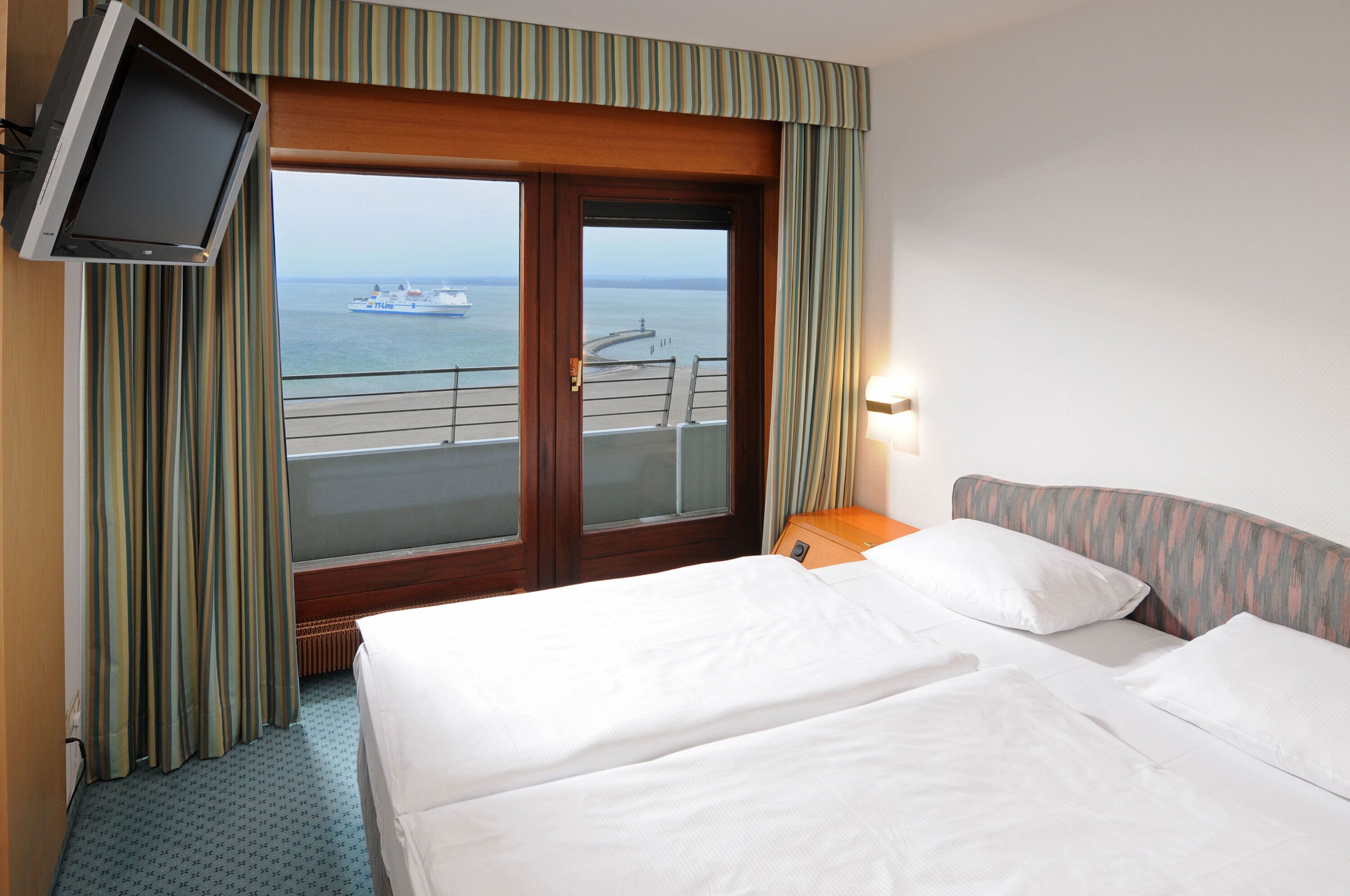 A hotel room with two beds, a television, and a view of the sea and a ship.