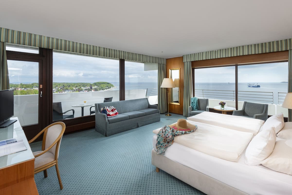 A hotel room with a large bed, a sofa, a desk with a computer, and a TV. There are large windows with a view of the sea and a cityscape.