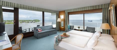 Habitación de hotel con una cama grande, un sofá, un escritorio con una computadora y un televisor. Amplias ventanas ofrecen vistas al mar y al paisaje urbano.