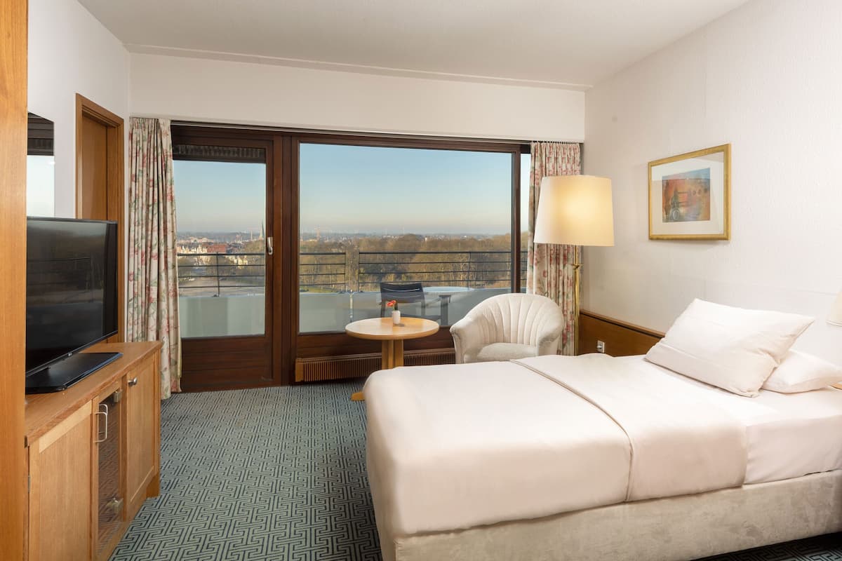 A hotel room with a bed, a TV, a desk, a chair, a window with a view, and a painting on the wall.