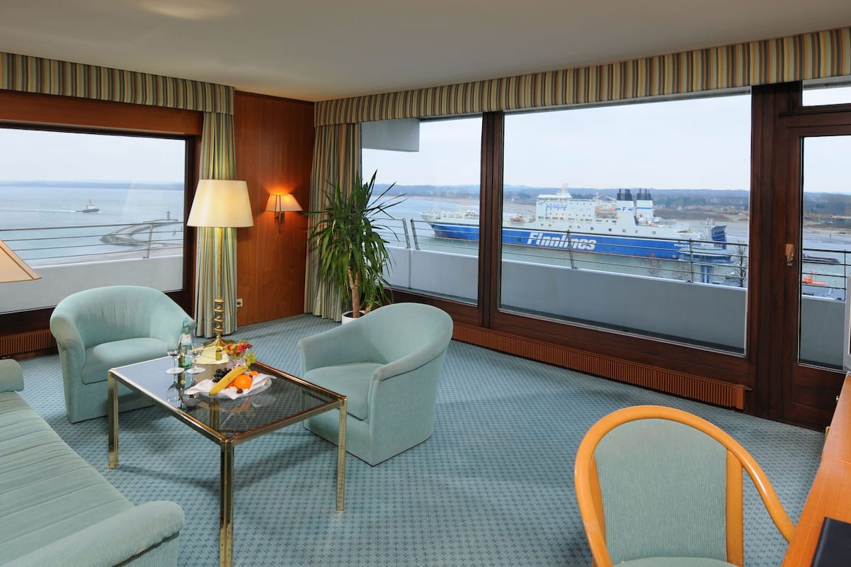 A hotel room with two armchairs, a small table with fruit, a lamp, and a view of a ship through large windows.