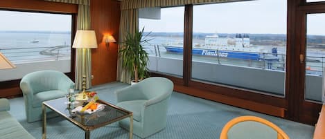 Habitación de hotel con dos sillones, una pequeña mesa con fruta, una lámpara y vistas a un barco a través de amplias ventanas.