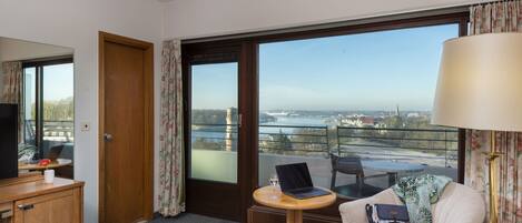 Habitación de hotel con una cama, un escritorio con una computadora portátil, una silla, una mesa con un vaso y vistas a un paisaje urbano a través de ventanas amplias.