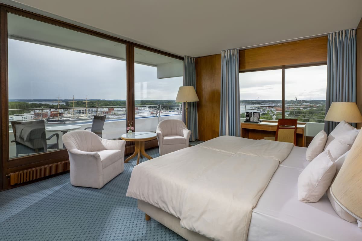 A hotel room with a large bed, two armchairs, a small round table, and a desk. The room has large windows offering a view of the city and a body of water.