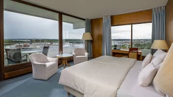Une chambre d’hôtel avec un grand lit, deux fauteuils, une petite table ronde et un bureau. La chambre dispose de grandes fenêtres offrant une vue sur la ville et sur un plan d’eau.