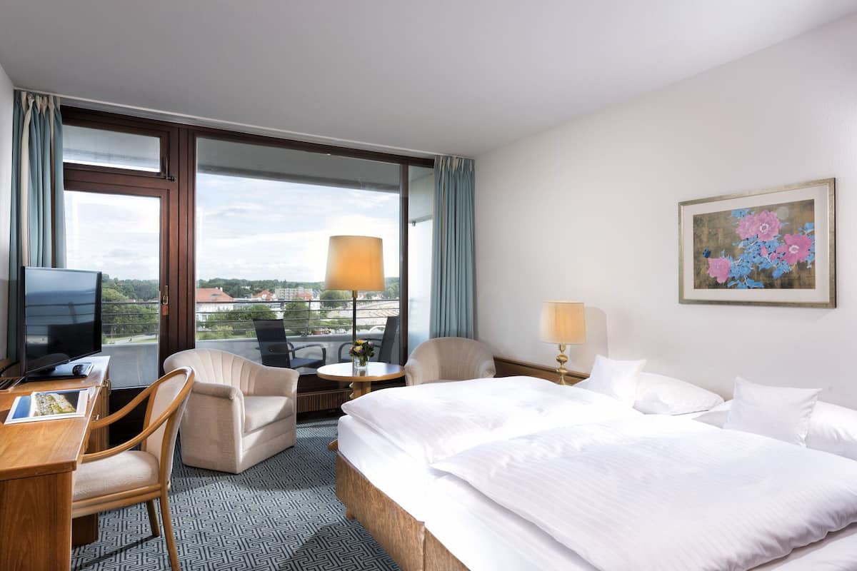 A hotel room with a bed, a desk with a computer, a chair, a small table with a lamp, and a painting on the wall.