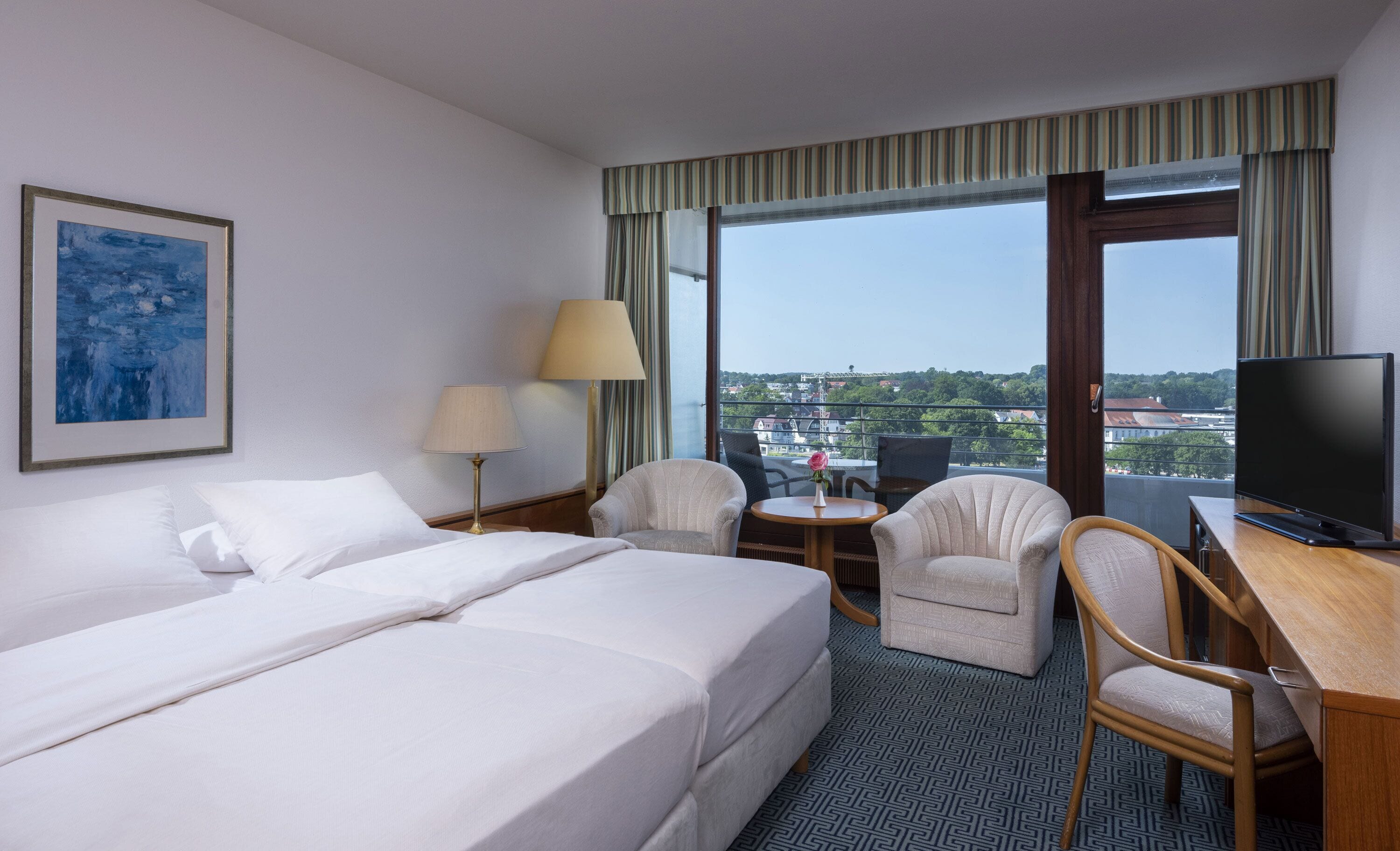 A hotel room with a bed, two chairs, a desk with a TV, a lamp, and a painting on the wall.