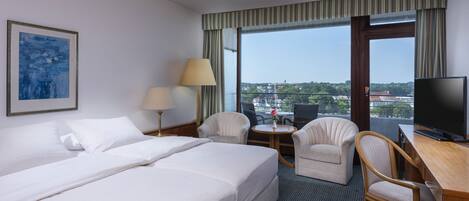 A hotel room with a bed, two chairs, a desk with a TV, a lamp, and a painting on the wall.